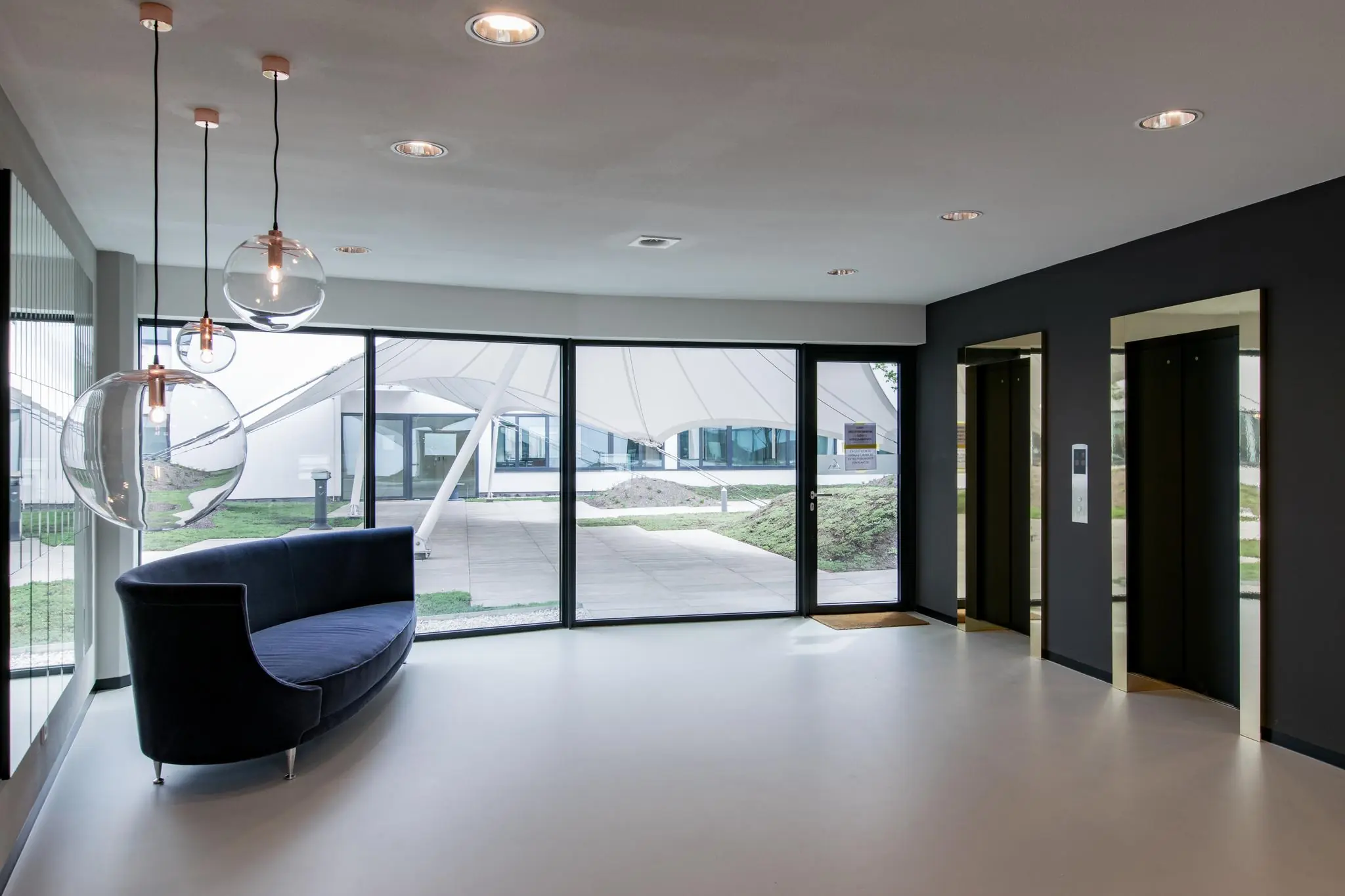 Moderne ontvangsthal met glazen hanglampen, een donkerblauwe bank, twee liften en grote ramen met uitzicht op een overkapt buitenterrein.